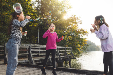 Wall Mural - Three girls blowing bubbles on a wooden dock by a lake during sunset. The scene captures their joyful expressions and the shimmering bubbles floating in the air.