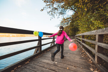 A young girl in a pink jacket runs along a wooden bridge by a lake, holding colorful balloons. The scene is set in a serene natural environment with trees and a clear sky.