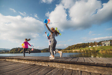 Wall Mural - Three girls joyfully walking along a wooden pier by a lake, each holding colorful balloons. The scene is bright and cheerful, with trees and mountains in the background.