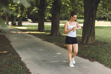 A young woman jogging in a park, checking her smartwatch. She wears a white tank top and black shorts, with a natural green background of trees and grass.