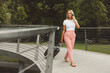 © VisualProduction - A woman walking on a curved bridge in a park, wearing a white t-shirt and pink pants, carrying a suitcase. The scene is serene with green trees in the background.