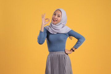 Wall Mural - happy asian indonesian muslim woman giving ok finger gesture on isolated yellow background