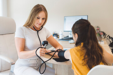 A young girl patient sitting at a table while a healthcare professional doctor measures her blood pressure. The girl looks calm and attentive, wearing a yellow top. The setting appears to be a medical