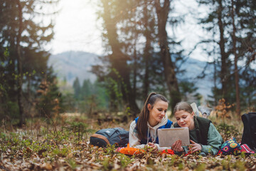 Two young women sitting on the ground in a forest, looking at a tablet together. They are surrounded by nature, with trees and mountains in the background. Both are smiling and engaged with the device