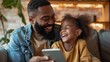 © Usman - A happy black father and daughter are sitting together on a couch at home, looking at a tablet. The father is smiling and holding the tablet, while the daughter is laughing and looking at the screen