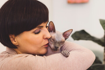 A woman with short dark hair gently kisses a Sphynx cat while holding it close. The cat appears relaxed and content in her embrace. The background features soft colors and greenery, creating a warm at