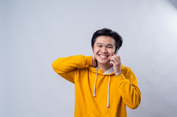 An Asian man in a yellow hoodie is happily talking on the phone. He has one hand behind his head, and he is smiling with eyes closed. The background is plain white