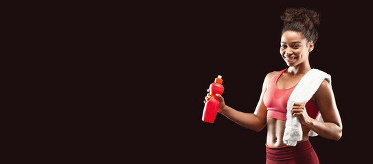 Wall Mural - Pretty smiling african sporty girl with towel on her shoulders drinking water, black studio background with copy space