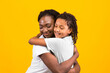 © Prostock-studio - Loving African Woman Embracing Her Child, Looking Happy And Calm. Yellow studio wall, copy space
