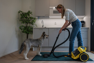 Young woman vacuuming carpet with her dog at kitchen