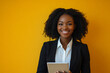 © Volodymyr - Young Black businesswoman holding a touchpad in an office, smiling brightly at the camera, against a yellow background.
