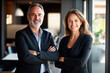 © GustavsMD - Smiling mature business professionals, a man and a woman, standing with arms crossed in a stylish office. Confident colleagues or partners in a corporate environment. Teamwork, leadership and business