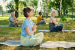 © pressmaster - Side view of serene mature woman in t-shirt and sweatpants sitting on mat in natural environment and practicing yoga exercises