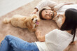 © Studio Romantic - Cheerful couple lying on floor, enjoying free time at home with their cute pet. Smiling wife and husband playing with maltipoo dog on carpet, petting and relaxing with puppy in loving atmosphere.