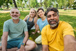 © pressmaster - Happy mature intercultural men and two women looking at camera while one of them holding smartphone and taking selfie in park