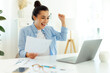 © Kateryna - Good job. Cheerful happy indian or arabian successful business woman, sitting at a workplace, joyfully looking at a laptop screen, rejoicing in success, holding documents, smiling, gesturing with fist