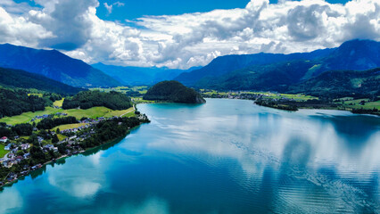 Aerial view of the serene Wolfgangsee in Austria, surrounded by green mountains and picturesque villages. The crystal-clear lake reflects the stunning alpine landscape, offering a peaceful and scenic 