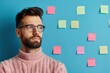© Vilaysack - A young man with glasses stands thoughtfully with crossed arms in front of a wall filled with colorful sticky notes, participating in a creative brainstorming session.