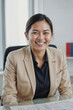 © polack - Asian manager woman in a business suit is smiling at the camera, she is sitting at a desk with a stack of papers in front of her. Concept of professionalism and confidence