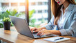 © PuriStudio - A woman is focused on her work while typing on laptop at wooden desk, surrounded by greenery. bright light enhances productive atmosphere