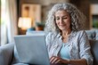 © ERiK - Happy curly middle aged woman using pc laptop surfing online sitting on sofa at home. Mature older lady looking away relaxing on couch with computer device in modern house living room
