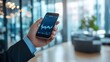 © Plaifah - A businessman holding a smartphone with a financial graph app open, with the focus on rising stock market trends, set against a clean office background.