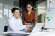 © Liubomir - Asian business professionals engaged in collaborative work in modern office setting. Man and woman discussing project details, using laptop and phone. Teamwork technology in professional environment