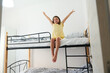 © Taras Grebinets - Joyful young girl in yellow dress sitting on upper bunk bed with arms raised in bright cozy bedroom