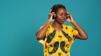 Wall Mural - Young adult having fun dancing and listening to her music in studio, enjoying a new playlist on audio headset against blue background. Showing groovy dance moves, traditional attire. Camera A.