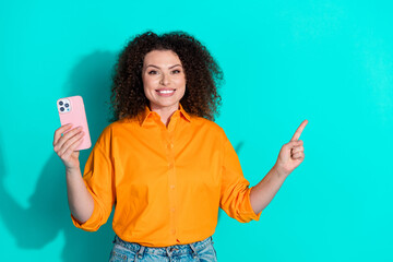 Wall Mural - Photo of nice smart woman with wavy hair dressed orange shirt hold smartphone directing empty space isolated on teal color background