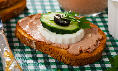 Wall Mural - Toast with meat pate and fresh cheese decorated with cucumber, black olive slice and greens on checkered textile background