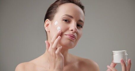 Beauty, health, cosmetics, anti-aging therapy and skin care concept - young beautiful brunette Caucasian woman holds a jar of moisturizer in her hands and applies the cream to her face skin