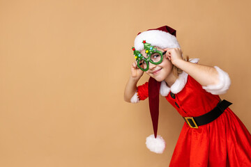 A cheerful child wearing a Santa costume and festive glasses poses joyfully against a warm, neutral backdrop during the holiday season