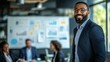 © arhendrix - Confident businessman with a charming smile, standing in a modern office with colleagues discussing in the background, in front of data charts.