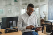 © mavoimages - Smiling African businessman leaning on an office desk using a tablet