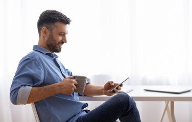 Poster - Break In Work. Relaxed Male Freelancer Using Smartphone And Drinking Coffee While Sitting At Desk At Home Office, Browsing Internet Or Social Networks, Enjoying Remote Job, Side View With Copy Space