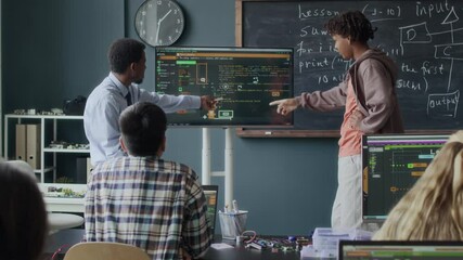 Medium shot of African American male teacher and multiethnic group of gen z students discussing software interface on monitor display during programming lesson in modern class