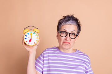 Sticker - Photo of sad upset mature woman wear striped stylish clothes hold alarm clock isolated on beige color background