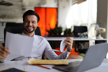 Wall Mural - A man is sitting at a desk with a laptop and a piece of paper. He is smiling and holding the paper in his hand. The scene suggests a positive and productive atmosphere