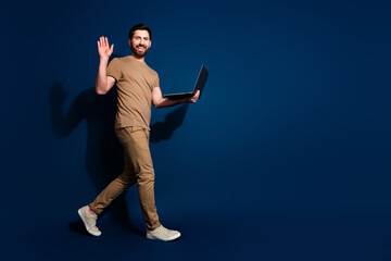 Wall Mural - Photo of mature age web 3 developer brunet man with laptop walking waving hand friendly isolated on dark blue color background