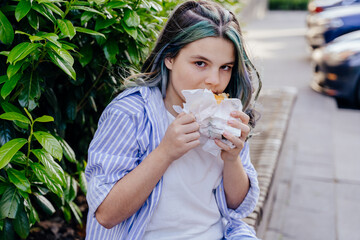 Hungry teenager girl with blue colorful trendy hairstyle biting crispy croissant outdoors on street. Close up.