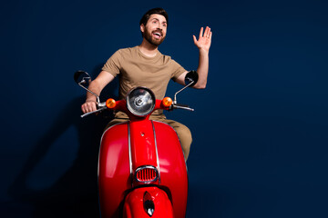 Canvas Print - Photo of happy young man in beige t shirt likes summertime riding motorbike waving hand deliver orders isolated on dark blue color background