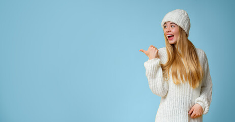 Cheerful girl in white knitted clothes pointing with thumb at empty space over yellow background