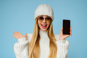 Wall Mural - Emotional woman in winter hat and sunglasses demonstrating blank smartphone screen over yellow background, free space