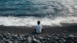 © TPS Studio - Man Sitting on Rocky Beach Facing Ocean Waves Under Cloudy Sky, Reflecting and Relaxing by the Seaside
