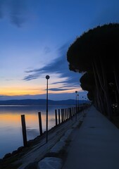 Poster - A quiet lakeside path lined with trees and lampposts, bathed in the fading light of dusk as the sky reflects over the calm waters below.