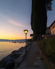 Poster - A tranquil lakeside walkway illuminated by warm lampposts, bordered by trees and rocks, as the sun sets, casting a soft glow over the water.