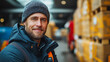 © CreativeIMGIdeas - A man wearing a black jacket and a black hat is smiling. He is standing in front of a warehouse with boxes