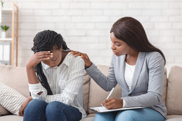 Female psychologist consoling depressed black woman at therapy session in office, supporting her to share problems