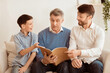 © Prostock-studio - Happy Grandfather Reading Book With Mature Son And Grandson Sitting On Couch At Home. Multigenerational Friendship. Selective Focus